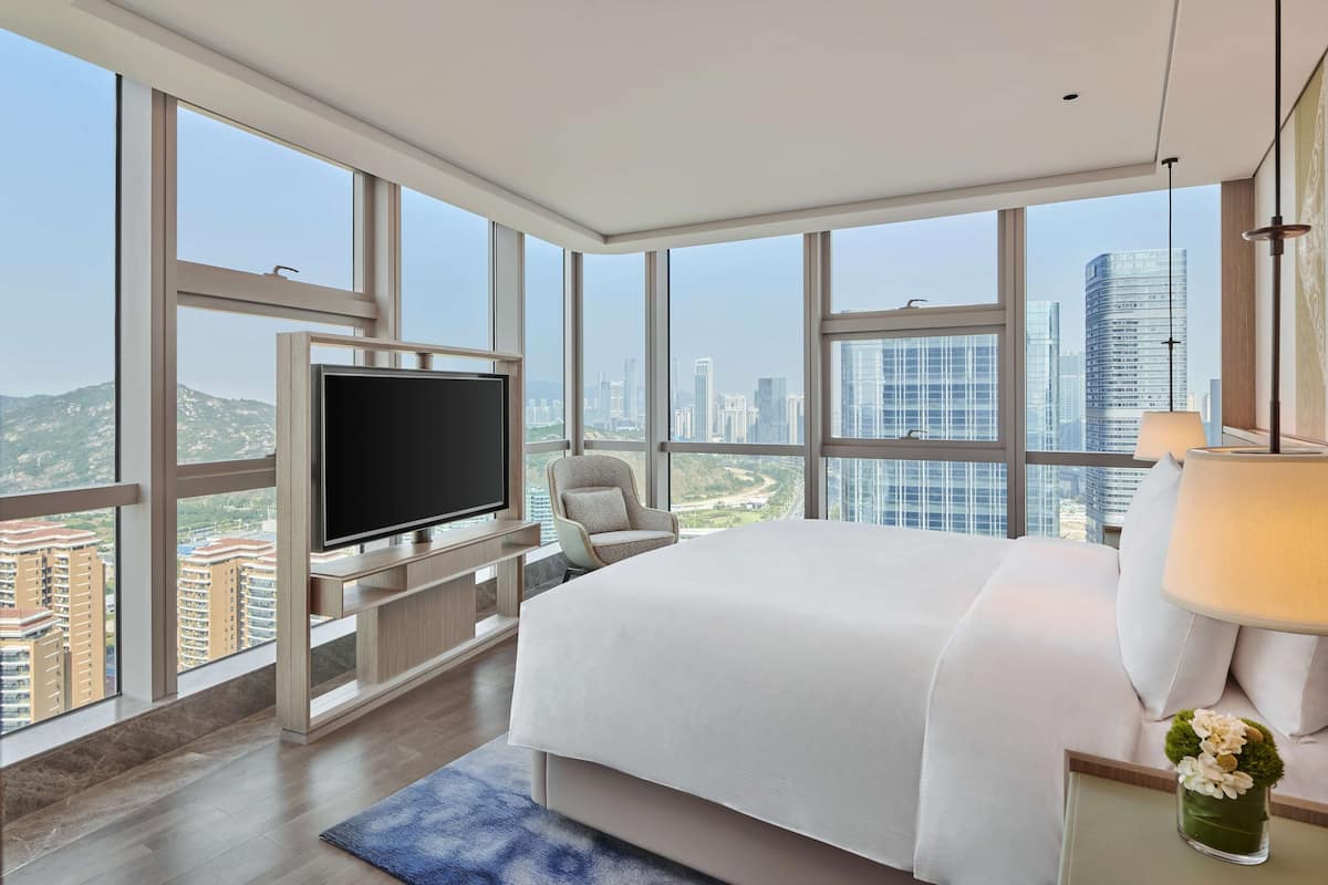 Una habitación de hotel moderna con una cama grande, un televisor de pantalla plana, una silla y vistas de la ciudad a través de amplios ventanales.