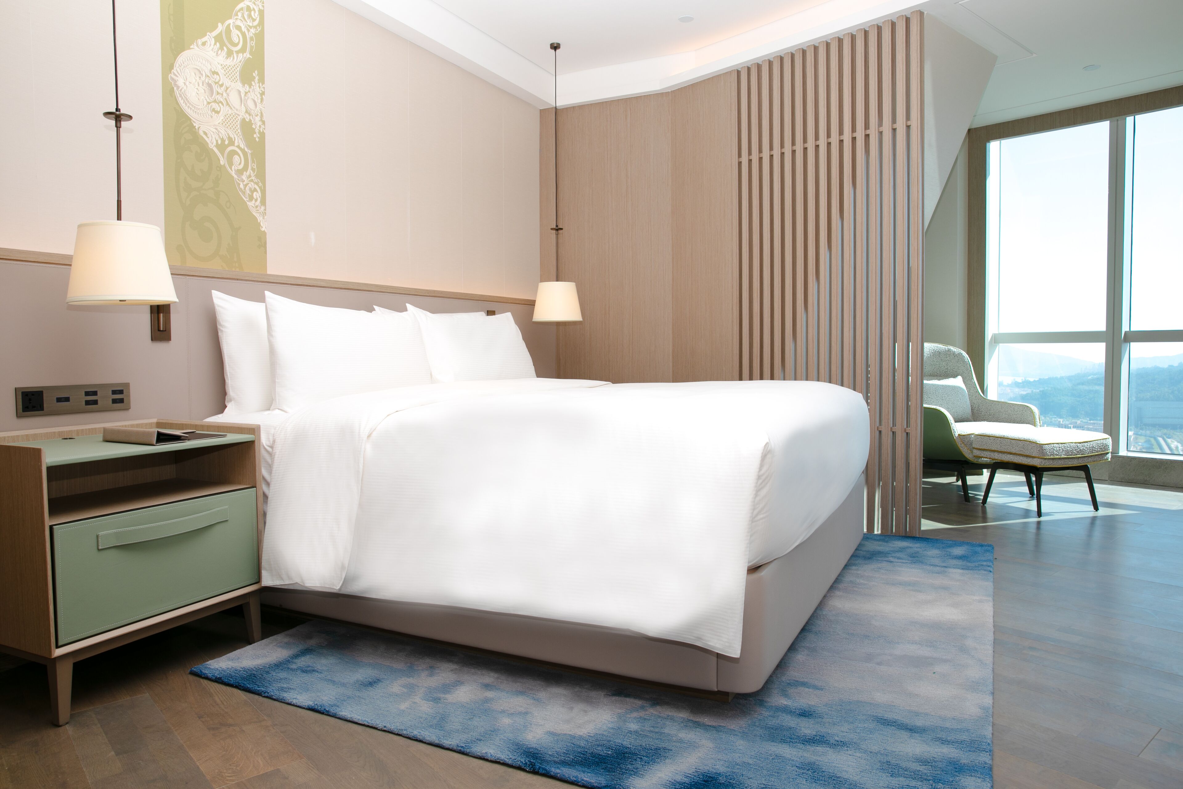 A modern hotel room with a large bed, a nightstand, a chair, and a view of the city.