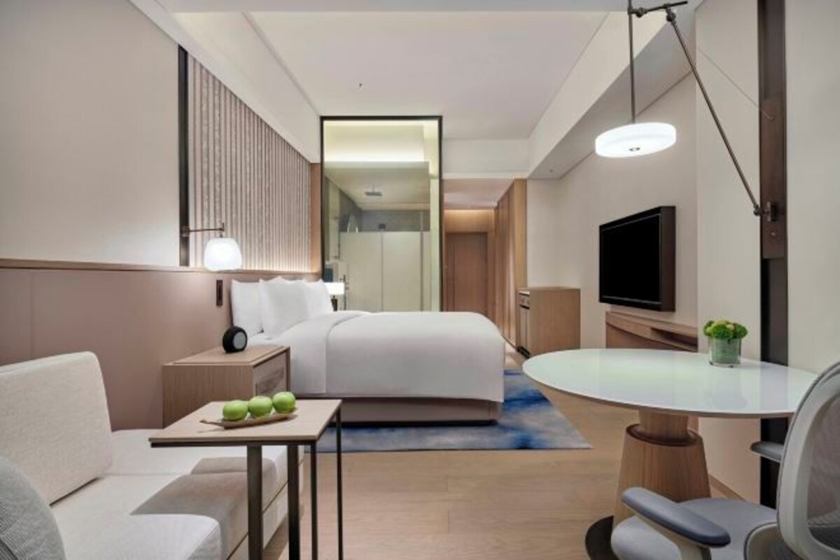 Une chambre d’hôtel moderne équipée d’un lit, d’une table de chevet, d’une lampe, d’une télévision et d’une petite table sur laquelle se trouvent des pommes.
