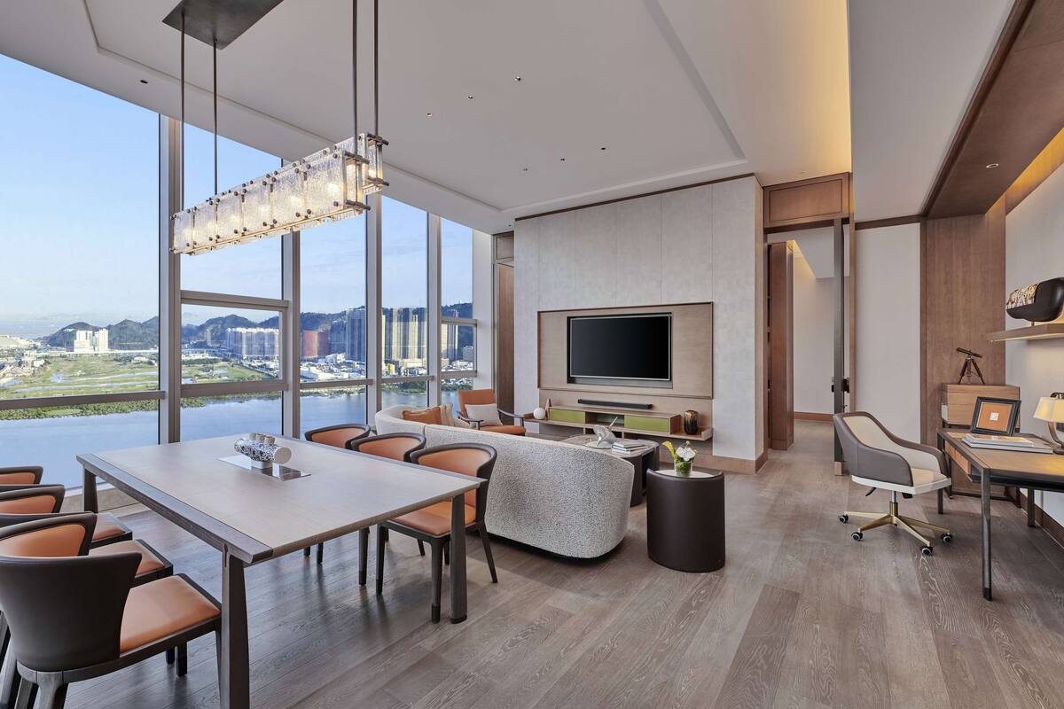 Una habitación de hotel moderna con un ventanal que ofrece vistas a la ciudad, un televisor de pantalla plana montado en la pared, una zona de comedor con mesa y sillas, y un rincón de estar con sofás y una mesa de centro.