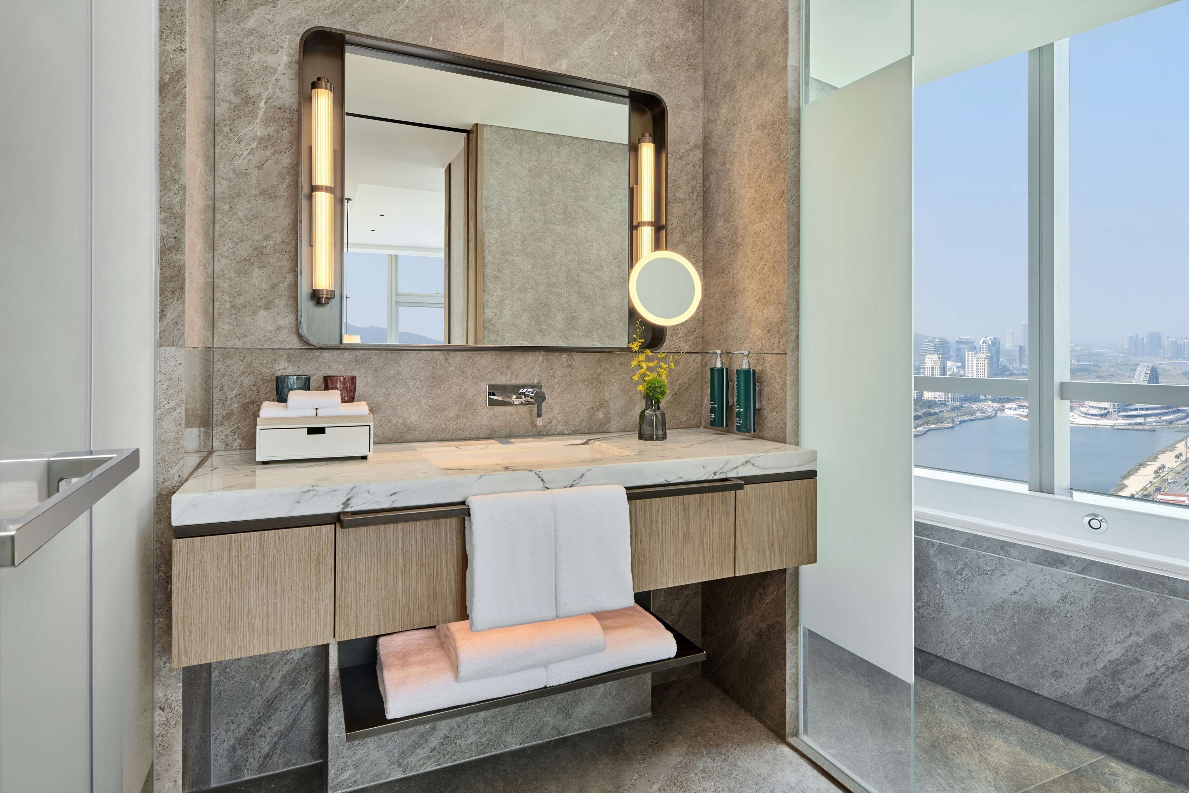 A modern bathroom with a large mirror, a vanity with a marble countertop, a built-in towel rack, and a view of a cityscape through a large window.