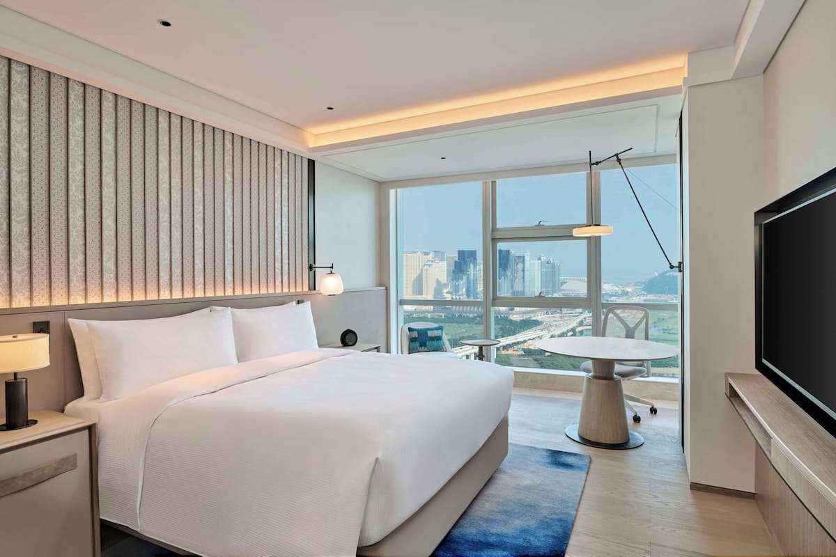 Un hotel moderno con una cama grande, televisor, escritorio con lámpara y vistas de la ciudad a través de amplias ventanas.