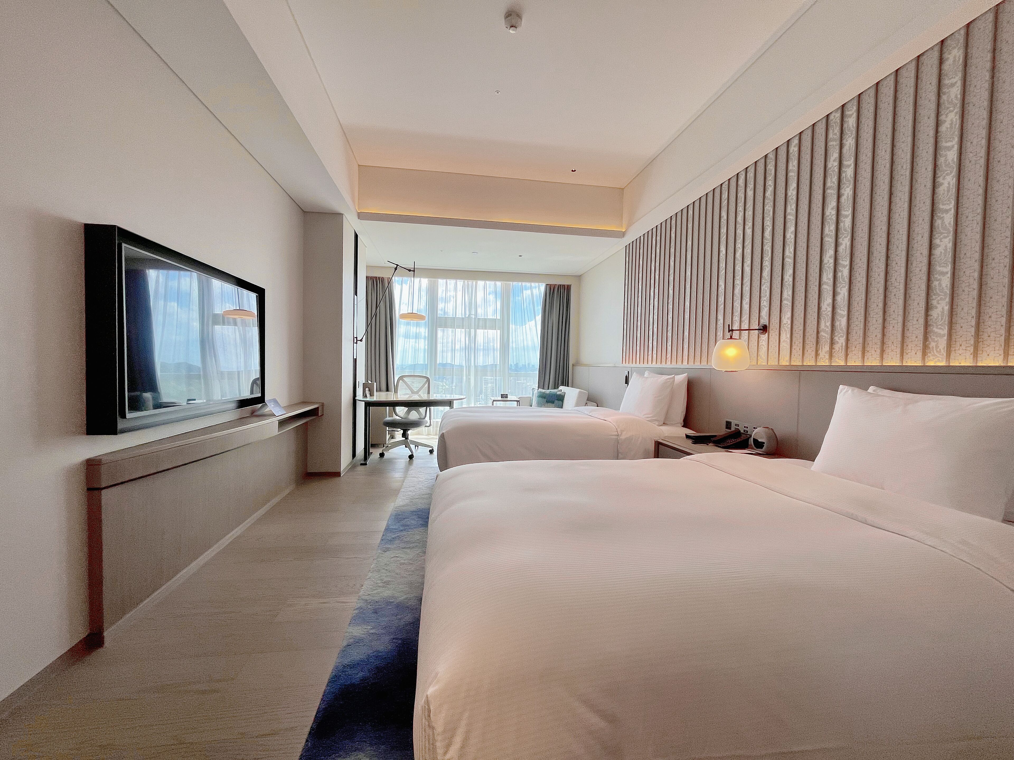 A hotel room with two beds, a desk, a TV, and a view of the city.