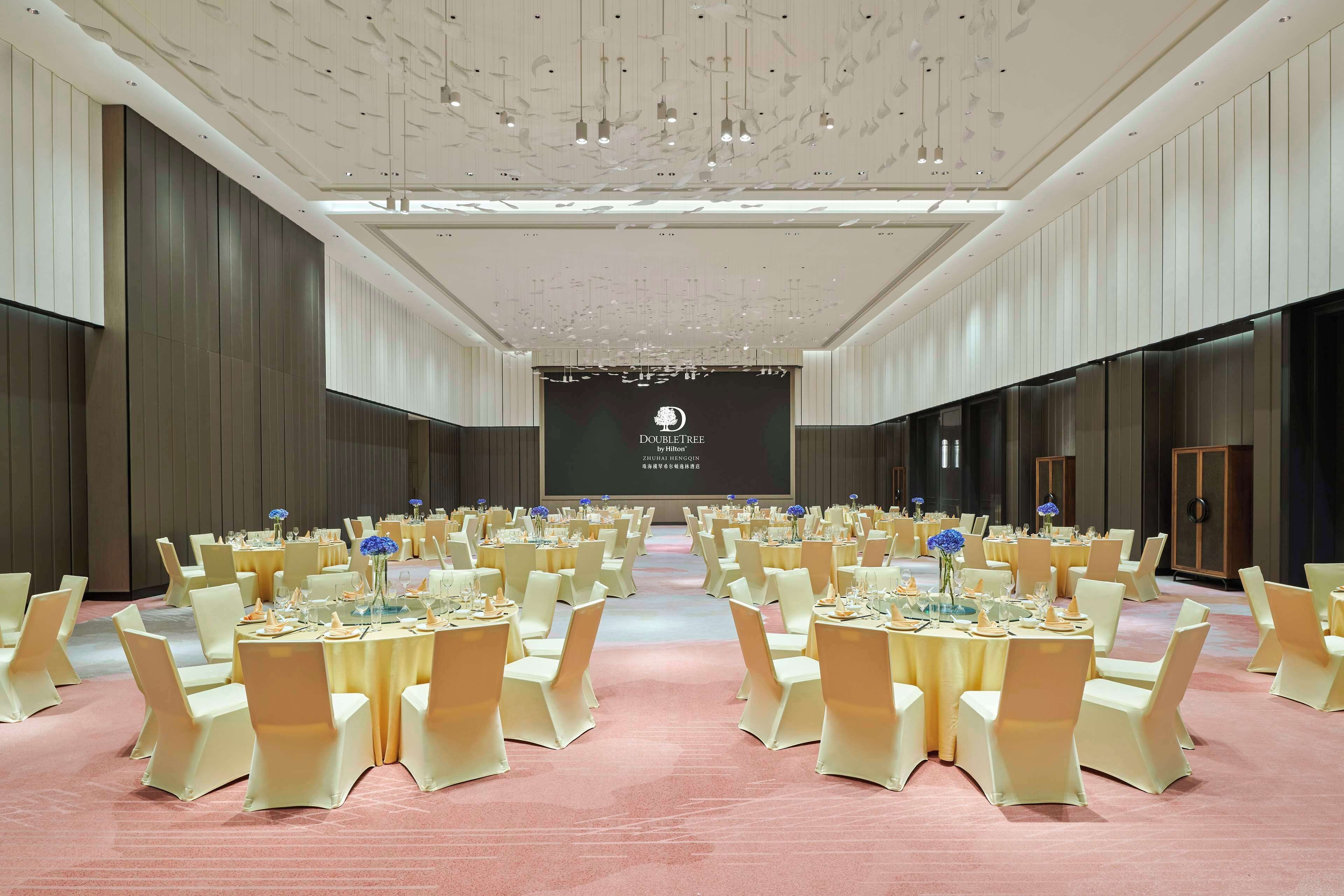 Une grande salle de banquet, bien éclairée, avec des rangées de tables et de chaises, une scène dotée d’un écran et un design de plafond moderne.