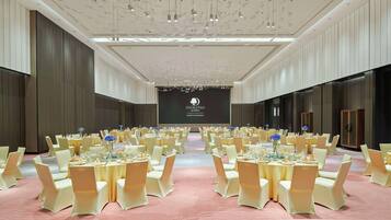 Une grande salle de banquet, bien éclairée, avec des rangées de tables et de chaises, une scène dotée d’un écran et un design de plafond moderne.