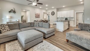 A modern living room with a sectional sofa, ottoman, and a dining area with a kitchen in the background. - Charming Ozark Abode w/ Yard & Pool Access (Ozark)