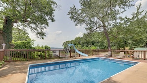 A rectangular swimming pool with a blue slide and a ladder, surrounded by a wooden deck and trees. - Large Lakefront Home | Pool, Hot Tub & Boat Dock (Grove)