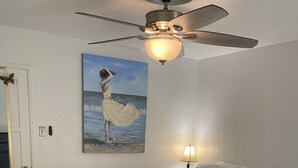 A bedroom with a bed, a ceiling fan, a painting of a woman by the sea, a lamp, and a door with a mirror. - Comfy Bungalow (Lone Pine)