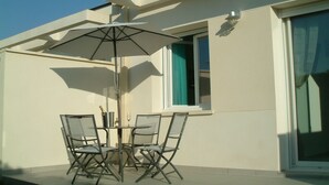 Une terrasse extérieure avec une table et des chaises, un grand parasol et une fenêtre avec un rideau.