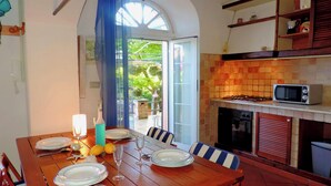 Un comedor con una mesa de madera preparada para el desayuno y la cena, una cocina con salpicadero de azulejos, y vista a un área exterior a través de una puerta de vidrio.