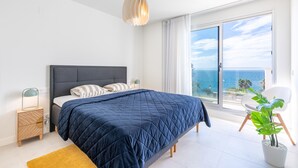 A bedroom with a large bed, a bedside table, a nightstand, a potted plant, and a view of the sea through a sliding glass door.