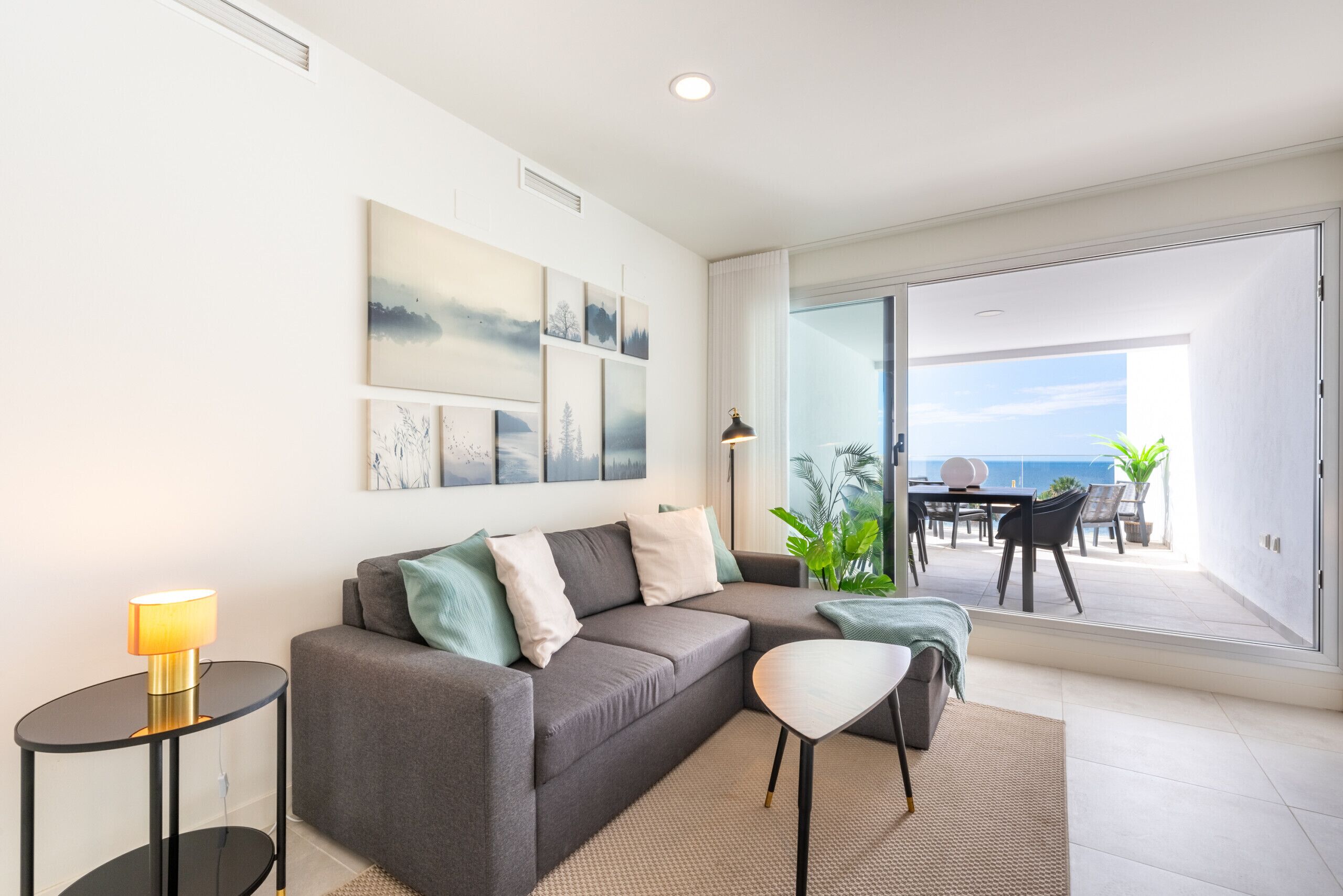 Een moderne woonkamer met een grijze bank, een klein bijzettafeltje met een lamp, en uitzicht op het landschap door een schuifdeur.