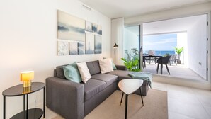 A modern living room with a grey sofa, a small side table with a lamp, and a view of the outdoors through a sliding glass door.
