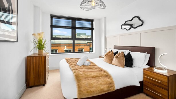 Une chambre à coucher moderne avec un grand lit, une table de chevet en bois, une lampe de chevet, une fenêtre avec des stores, un luminaire suspendu et un tableau encadré au mur.