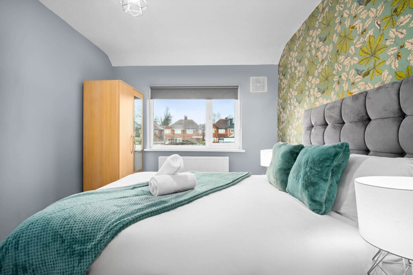 A modern bedroom with a large bed, a wooden wardrobe, a window with a view of houses, and a patterned wall.