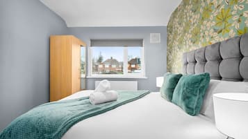 A modern bedroom with a large bed, a wooden wardrobe, a window with a view of houses, and a patterned wall.