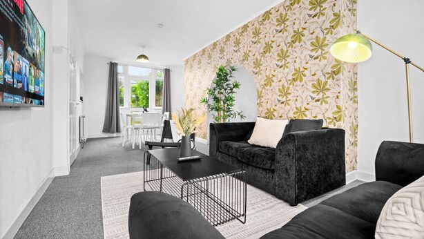 Ein modernes Wohnzimmer mit einem schwarzen Sofa, einem Couchtisch und einer Stehlampe. Im Hintergrund befindet sich ein Essbereich mit Tisch und Stühlen.