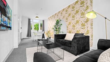 A modern living room with a black sofa, a coffee table, and a floor lamp. There is a dining area with a table and chairs in the background.