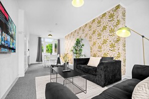 Un salon moderne avec un canapé noir, une table basse et un lampadaire. On aperçoit à l’arrière-plan un coin repas avec une table et des chaises.