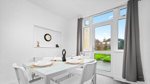 Ein Essbereich mit einem weißen Tisch, gedeckt für vier Personen, einer Uhr an der Wand und Blick auf einen Garten durch ein großes Fenster.