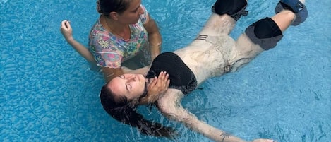 A person floating in a clear blue pool with another person standing beside them.