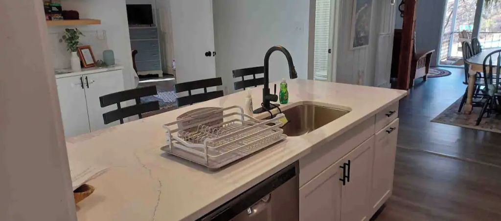 A kitchen with a white countertop, a sink, and a dish rack. There is a window with blinds and a door leading to another room.