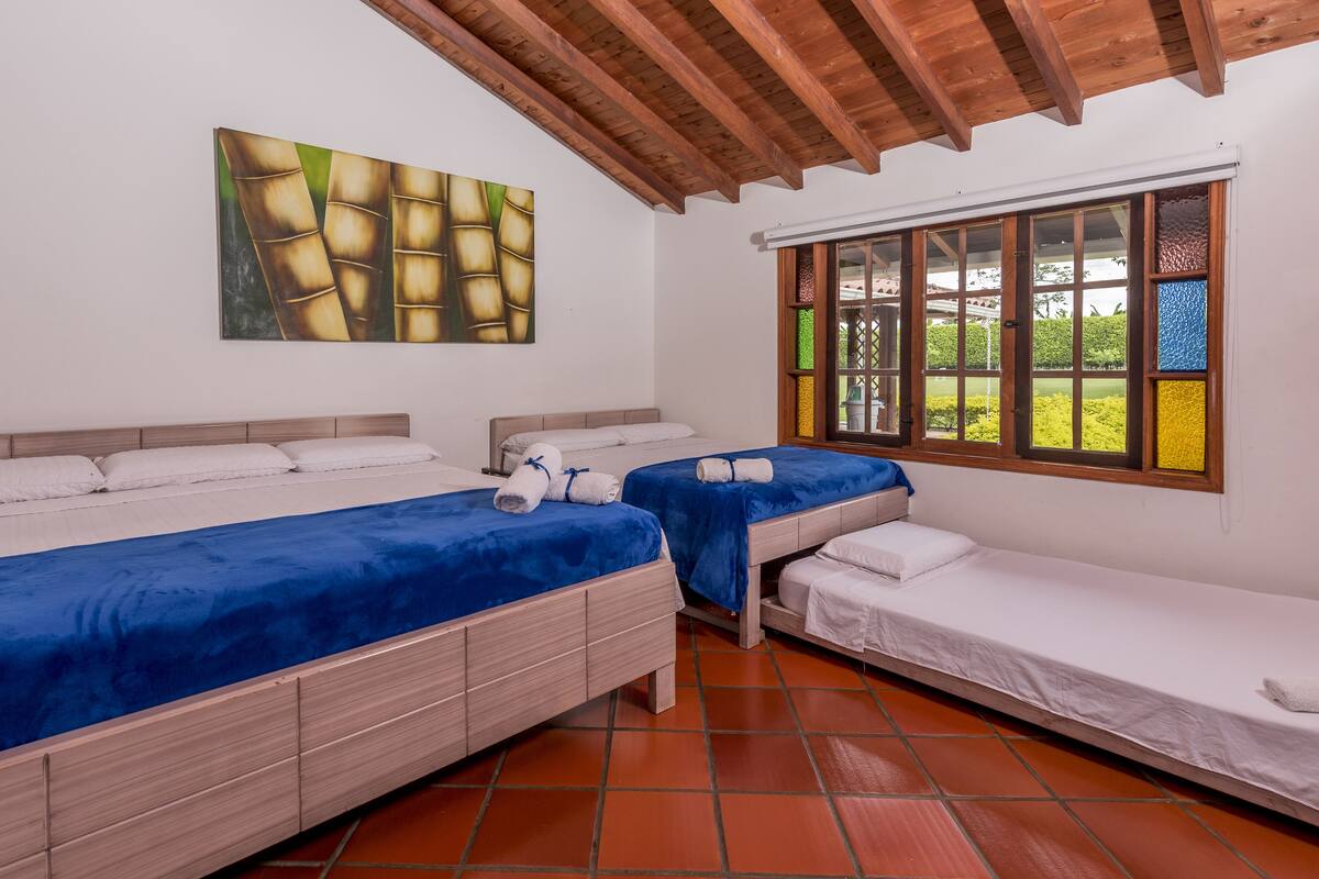 Ein Zimmer mit zwei Betten, eines mit blauer Bettdecke und das andere mit weißer Bettdecke. An der Wand hängt ein Gemälde und ein Fenster bietet Blick auf Grünflächen.