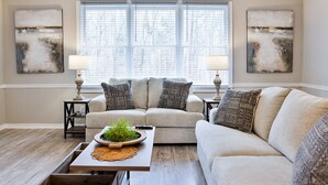 A living room with a beige sofa, two side tables with lamps, a coffee table with a plant, and abstract wall art.