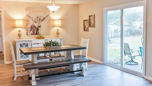 A dining area with a wooden table, white chairs, and a bench. There are two wall-mounted lamps, a chandelier, and framed artwork.