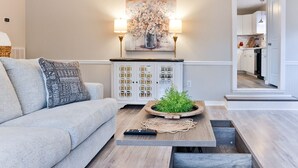 A living room with a grey sofa, a wooden coffee table with a plant, a wall-mounted painting, and a kitchen visible through an open door.