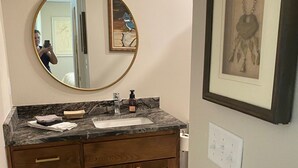 Une salle de bain avec un meuble-lavabo en bois, un miroir rond, une œuvre d’art encadrée et un luminaire placé au-dessus du miroir.