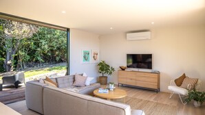 A modern living room with a large sofa, a coffee table, a TV, and a view of the outdoors. - Wavesong Whakaaio Onetangi | Be My Guest (Waiheke Island)