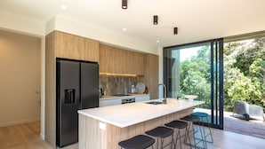 A modern kitchen with a large island, bar stools, a built-in refrigerator, and a view of the outdoors. - Wavesong Whakaaio Onetangi | Be My Guest (Waiheke Island)