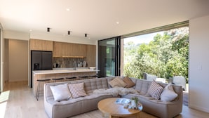 A modern living room with a large sectional sofa, a wooden coffee table, and a view of a kitchen and outdoor greenery. - Wavesong Whakaaio Onetangi | Be My Guest (Waiheke Island)