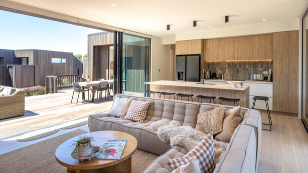 A modern living room with a sofa, coffee table, and a dining area with chairs. - Wavesong Whakaaio Onetangi | Be My Guest (Waiheke Island)
