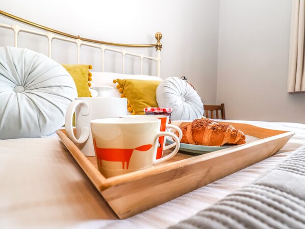 A neatly made bed with pillows, a tray with a croissant and mugs, and a bedside table with a lamp.
