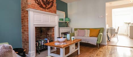 A cozy living room with a brick fireplace, a green sofa, a wooden coffee table, and a kitchen visible through an open doorway.
