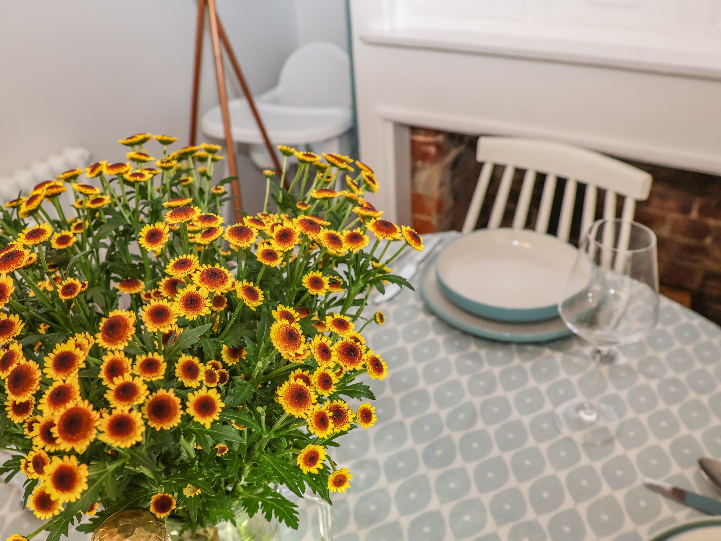 Una mesa con mantel estampado, un florero con flores amarillas y una silla blanca.