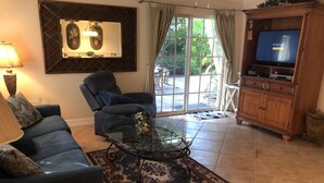 A living room with a sofa, armchair, glass coffee table, and a TV on a wooden cabinet. - Villa-style condo with pool on the beach - Condo 1 (Holmes Beach)