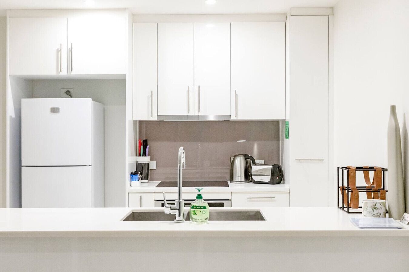 A modern kitchen with white cabinets, a built-in oven, a sink with a faucet, and a coffee maker.