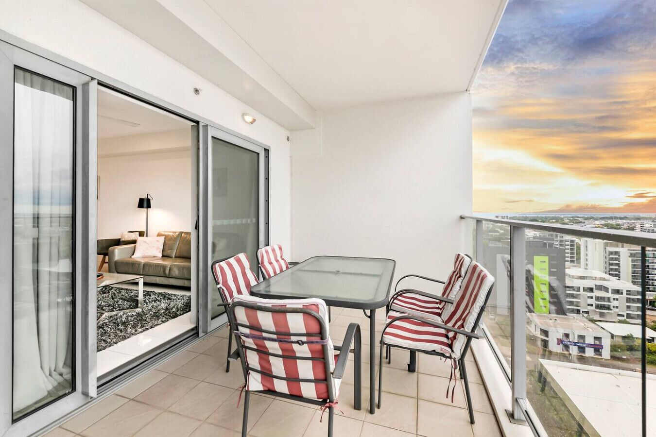 Un balcon avec une porte-fenêtre donnant sur un espace de vie, une table avec des chaises et une vue sur la ville au coucher du soleil.