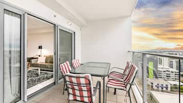 Un balcon avec une porte-fenêtre donnant sur un espace de vie, une table avec des chaises et une vue sur la ville au coucher du soleil.