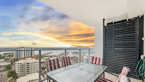 Een balkon met een glazen tafel en gestreepte stoelen, met uitzicht op een stadsbeeld en een zonsondergang.