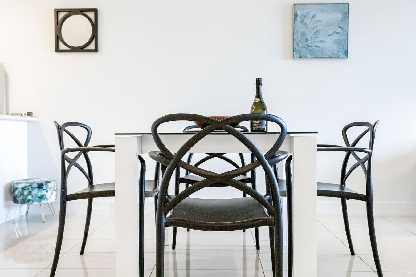 A dining area with a white table and four chairs with a crisscross design. A bottle of wine is on the table.