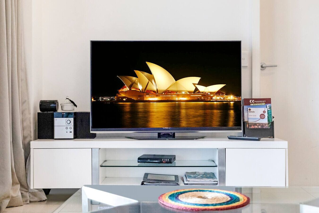 A flat-screen TV on a white entertainment unit with a Sydney Opera House image on the screen, a remote control, a book, and a glass coffee table with a colorful coaster.