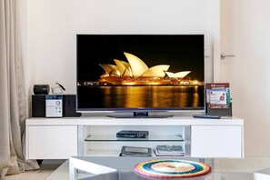 Un téléviseur à écran plat posé sur un meuble blanc, l’écran affichant une image de l’Opéra de Sydney, une télécommande, un livre et une table basse en verre surmontée d’un dessous de verre coloré.