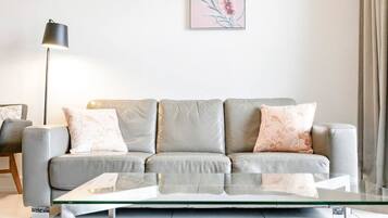 A modern living room with a grey sofa, two patterned cushions, a glass coffee table, a floor lamp, and a framed floral artwork on the wall.