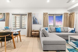 A modern living room with a grey sectional sofa, a dining table with chairs, and large windows with patterned curtains. - Reef-side Splendor: City Vistas ~ Pool ~ Workspace (Darwin City)