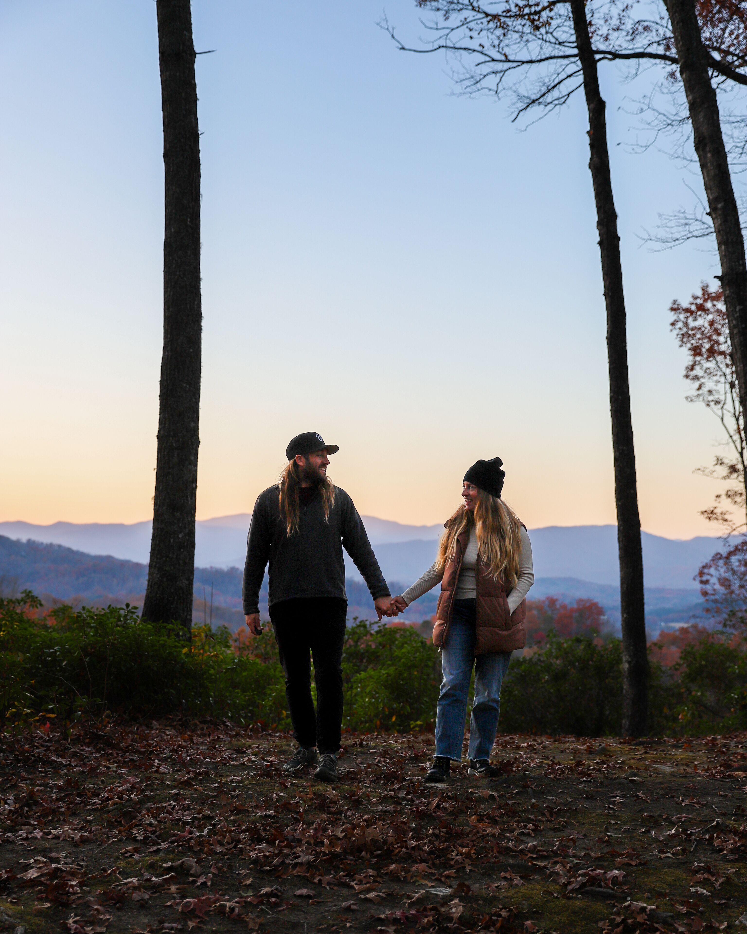 Two people holding hands on a leaf-covered path with trees and a distant mountain range.