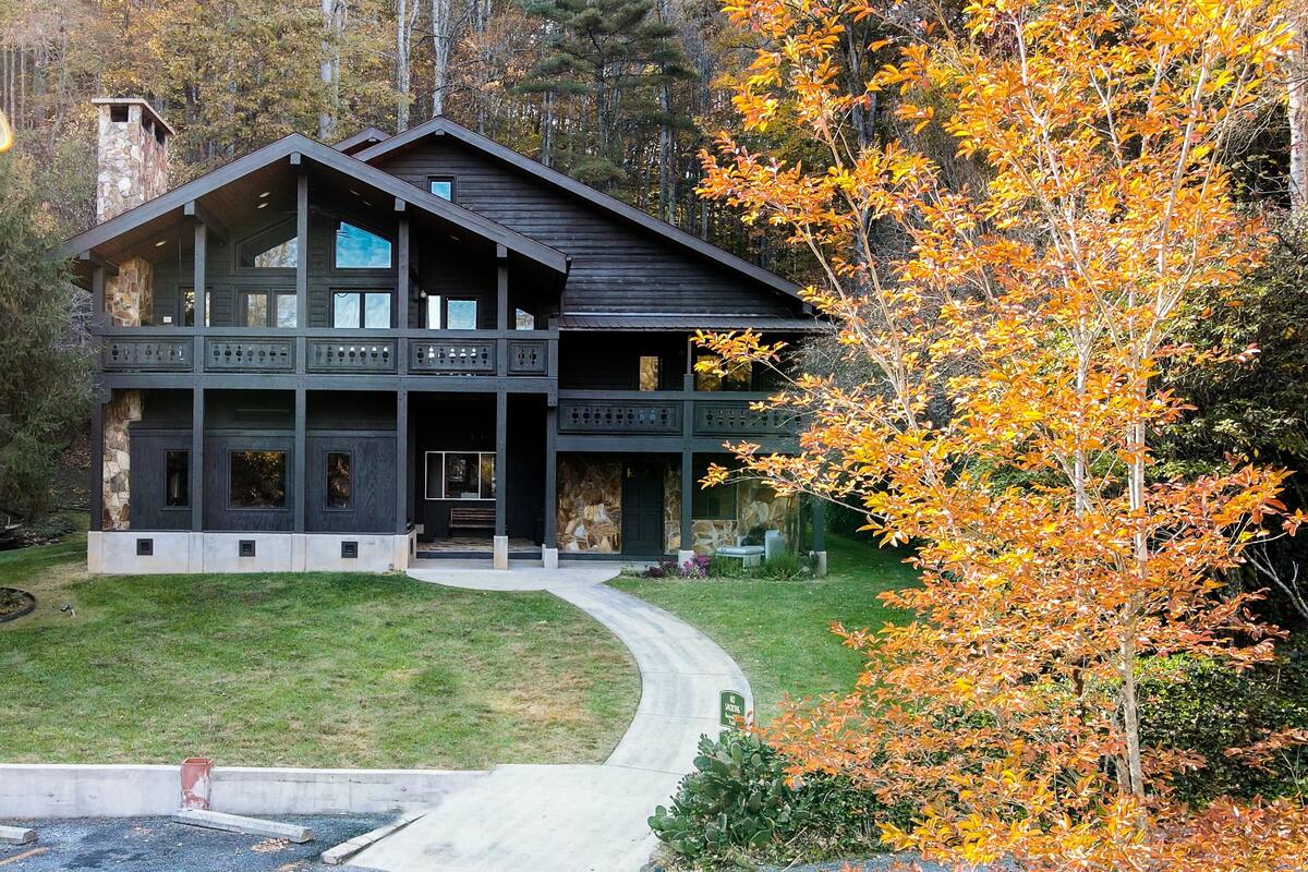 Eine große, dunkle Holzhütte mit mehreren Ebenen und Balkonen, umgeben von einem gepflegten Rasen und herbstlich gefärbten Bäumen.